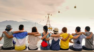 Eight friends sit shoulder to shoulder, arms around one another's shoulders, while the sit on a wall over looking the sea and a mountain, aerial tram crams move across the sky in the upper right corner.